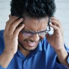 A man is holding his head and making a painful expression. He is wearing a blue shirt and black-framed glasses. 