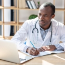 Doctor taking notes and smiling while looking at a laptop