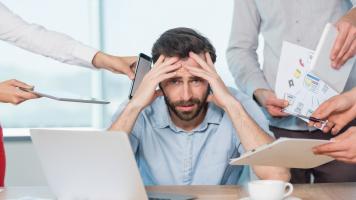Frustrated man surrounded by technology, papers, people.
