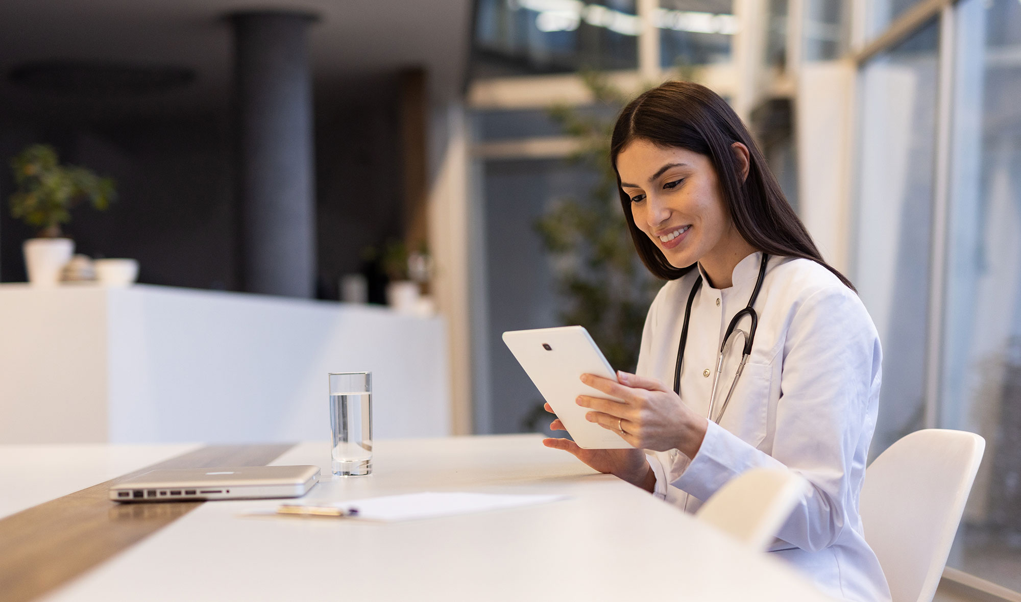 Healthcare professional in a white coat using a tablet at a desk.