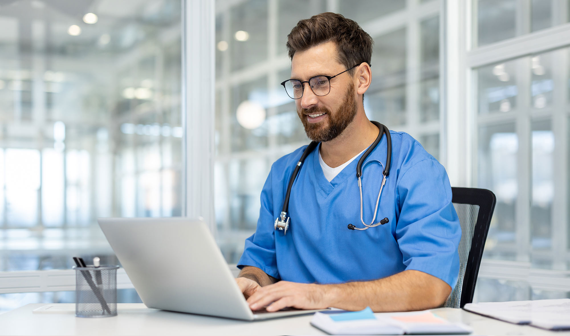 Healthcare professional in blue scrubs using a laptop at a desk.