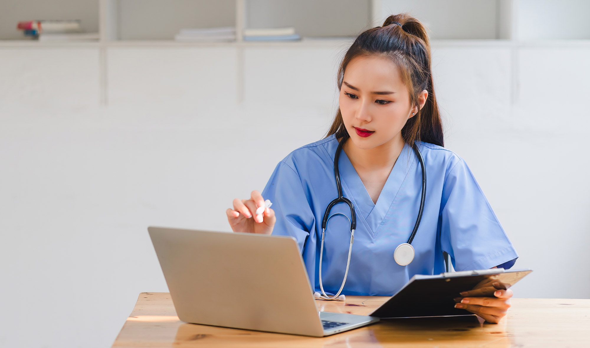 Healthcare professional in blue scrubs using a laptop and tablet at a desk.