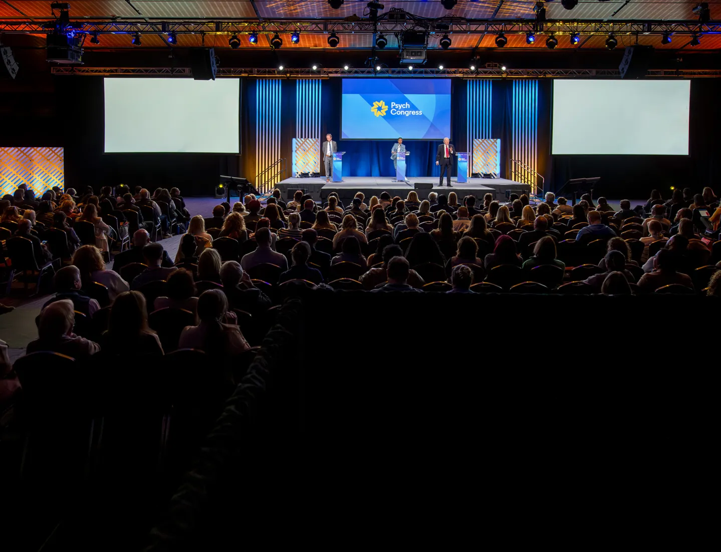 Full conference room watching three speakers on the main stage at Psych Congress