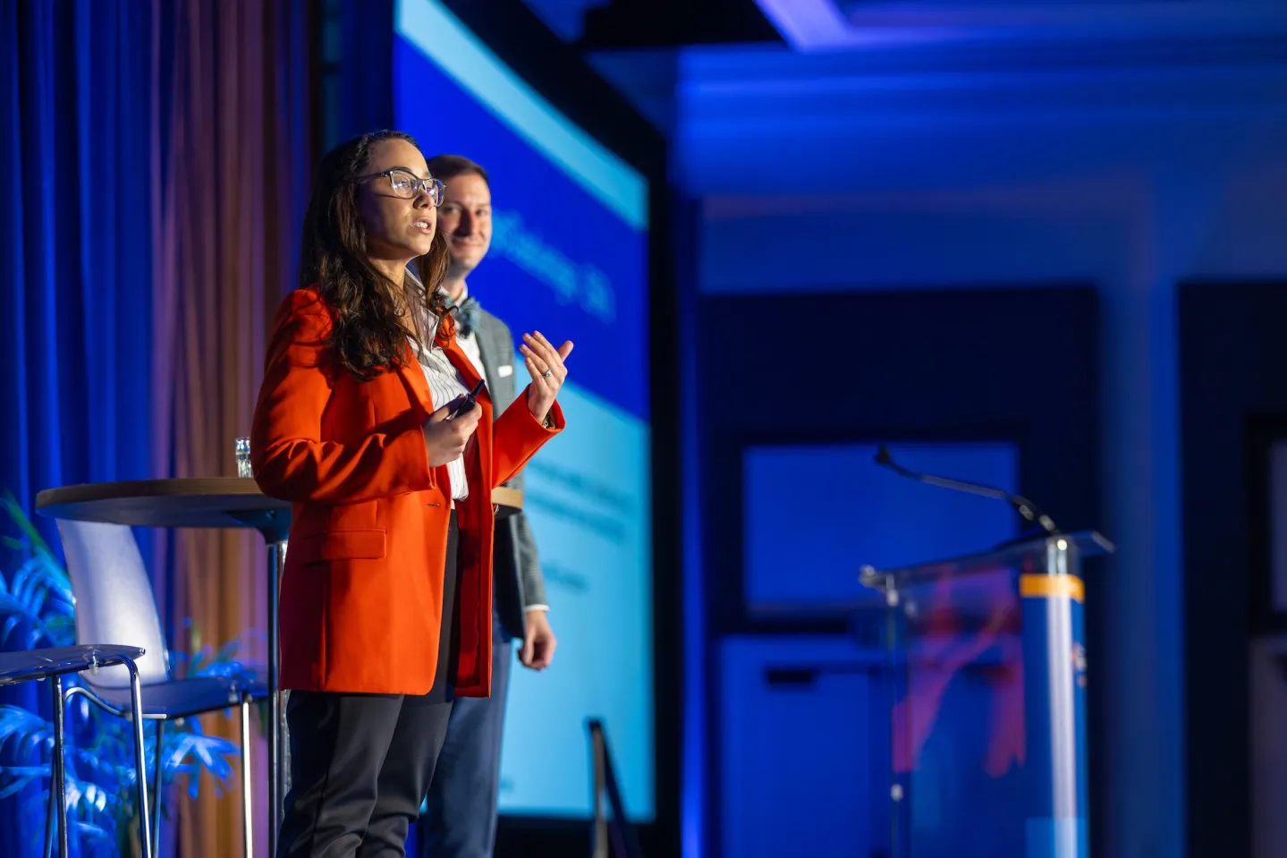 Woman in an orange jacket addresses an audience at a conference