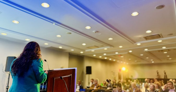 Speaker at a podium bathed in blue and yellow light speaking out to the full audience