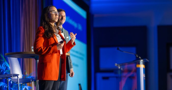 Woman in an orange jacket addresses an audience at a conference